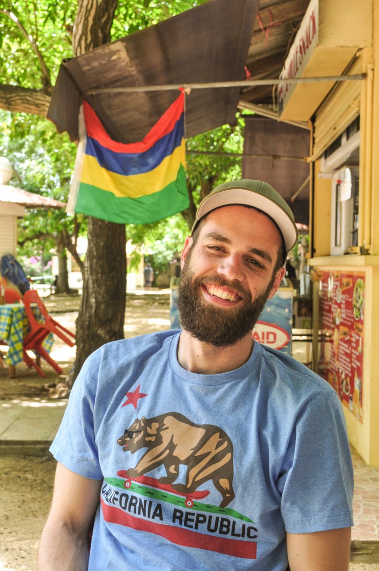 A young bearded man smiles in front of a Mauritian flag