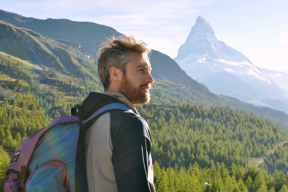A man with a backpack looks at a forested scenery featuring the Matterhorn