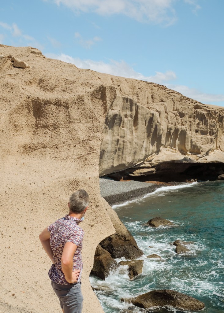Man looking at a pebbled beach beneath sandstone cliffs