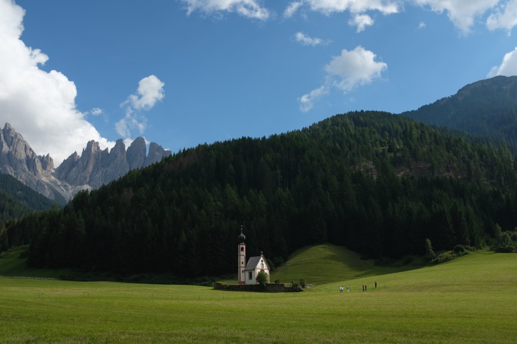 A small church in a green field next to forests and mountains